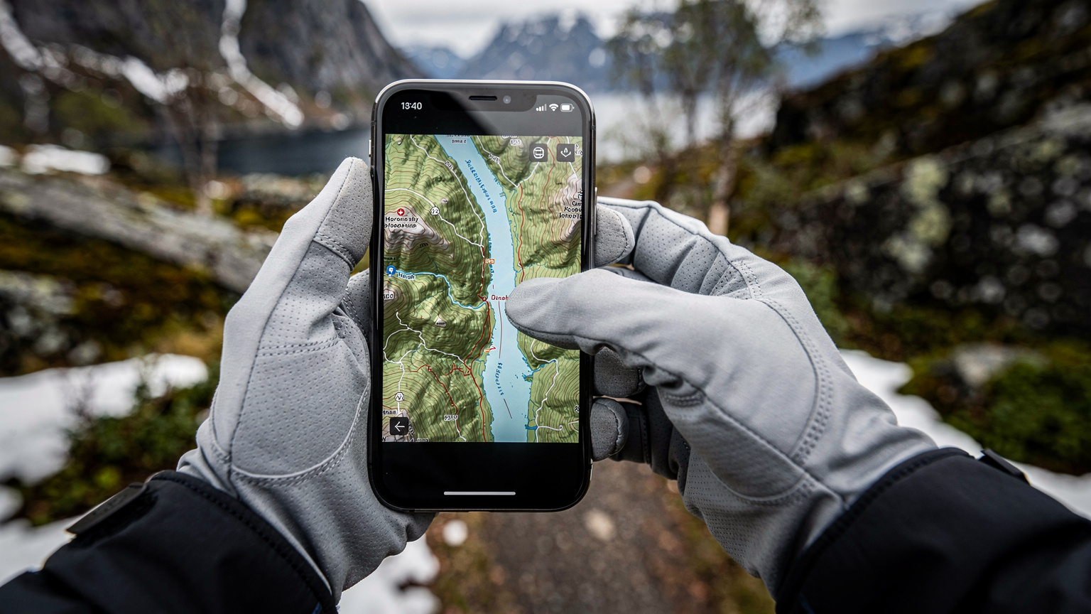 Close-up of a person's hands wearing light hiking gloves, holding a smartphone, with fingers navigating a map application showing a detailed topographic view of a Norwegian fjord, against a blurred background of a hiking trail.