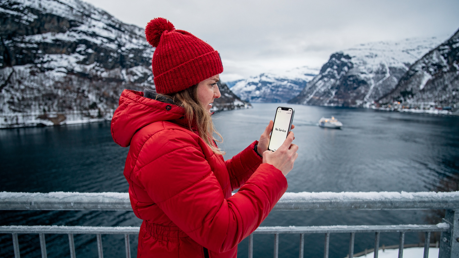 A traveler in a warm winter jacket looks confusedly at their smartphone screen, which shows a 'No Service' message, against the backdrop of a dramatic snowy fjord landscape, a ferry barely visible in the distance.