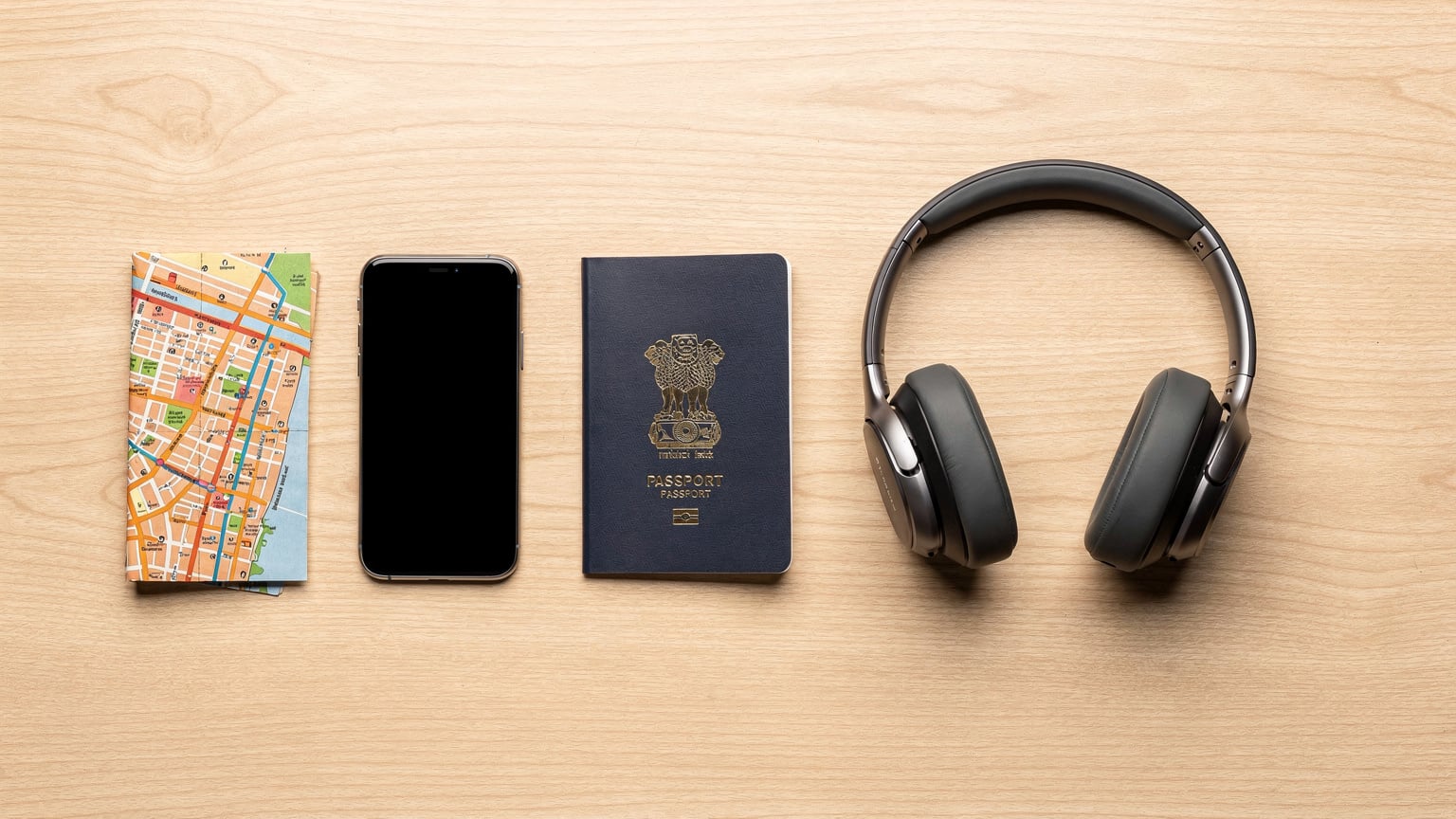 एक व्यवस्थित डेस्क पर एक पासपोर्ट, फोन, हेडफ़ोन और एक मुड़ा हुआ स्थानीय नक्शा रखा है।