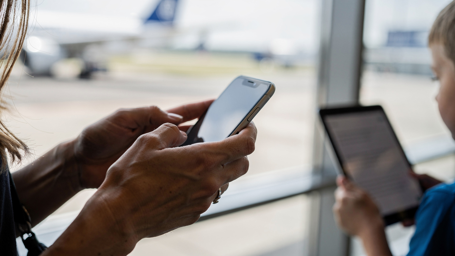 Bir annenin eli, Frankfurt Havalimanı'nda, diğer eliyle çocuğunun tabletine hotspot bağlantısı kurmak için telefonunda ayar yaparken görülüyor. Arka planda uçaklar bulanık bir şekilde geçiyor.
